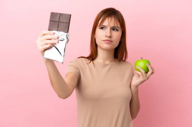 Kızıl saçlı kız pembe arka planda izole edilmiş, bir elinde çikolata tableti diğer elinde elma tutarken kuşkuları var.