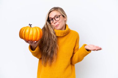 Beyaz arka planda izole edilmiş bir balkabağı tutan genç sarışın kadın omuzlarını kaldırırken şüpheyle hareket ediyor.