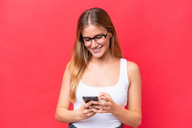 Kırmızı arka planda izole edilmiş genç ve güzel bir kadın cep telefonuyla mesaj gönderiyor.