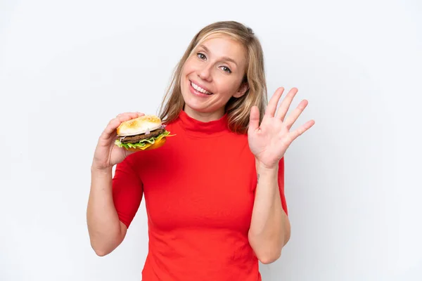 Młoda Blondynka Trzyma Hamburgera Białym Tle Salutując Ręką Szczęśliwym Wyrazem — Zdjęcie stockowe