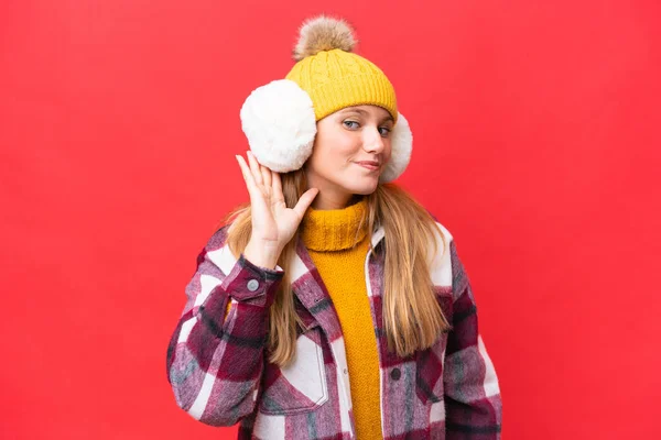 耳に手を置くことによって何かを聞く赤の背景に隔離された冬のマフを身に着けている若い美しい女性 — ストック写真