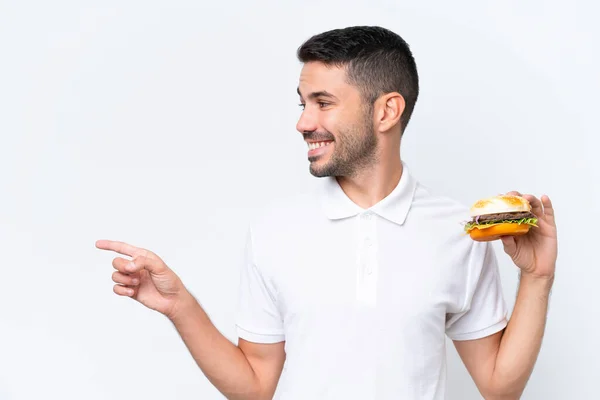 Młody Przystojny Biały Mężczyzna Trzyma Hamburgera Odosobnionym Tle Wskazując Bok — Zdjęcie stockowe