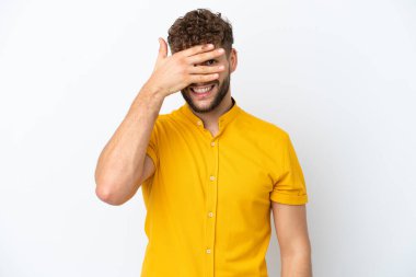 Beyaz arka planda soyutlanmış beyaz tenli yakışıklı bir adam. Gözleri elleriyle kaplıyor ve gülümsüyor.