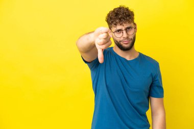 Sarı arka planda izole edilmiş genç, yakışıklı beyaz adam, olumsuz bir ifadeyle baş parmağını aşağı gösteriyor.