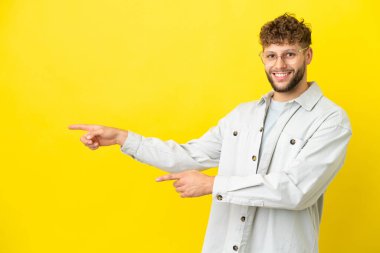 Sarı arka planda izole edilmiş genç, yakışıklı beyaz bir adam parmağını yana uzattı ve bir ürün sundu.