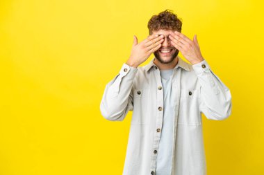 Sarı arka planda izole edilmiş yakışıklı beyaz bir adam. Gözleri elleriyle kaplıyor ve gülümsüyor.