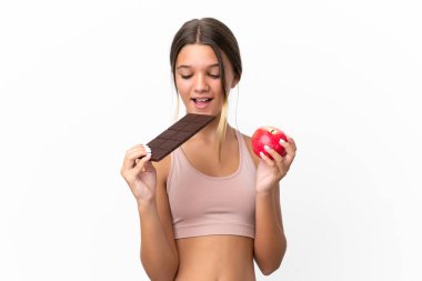 Beyaz arka planda izole edilmiş küçük beyaz kız bir elinde çikolata tableti, diğerinde elma alıyor.