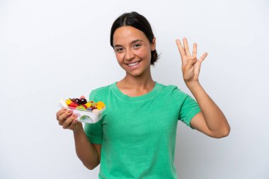 Beyaz arka planda bir kase meyve tutan genç İspanyol kadın mutlu ve parmaklarıyla dört tane sayıyor.