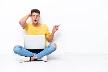 Genç adam beyaz arka planda izole bir şekilde yerde oturuyor. Şaşırmış ve işaret ediyor.