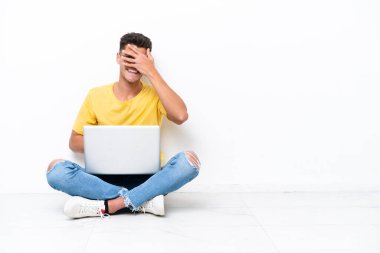 Genç adam, beyaz arka planda izole bir şekilde yerde oturuyor. Gözleri elleriyle kaplıyor ve gülümsüyor.