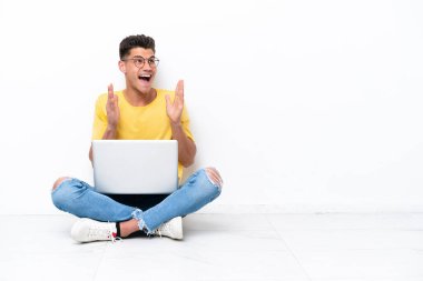 Yerde oturan genç adam, beyaz arka planda, şaşırmış bir yüz ifadesiyle izole edilmiş.