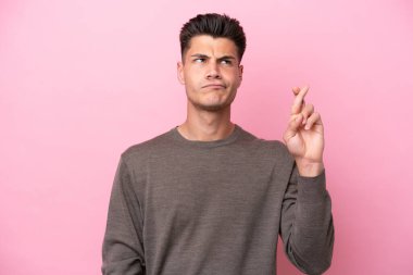 Pembe arka planda izole edilmiş genç beyaz adam parmaklarını çapraz yapıp en iyisini diliyor.