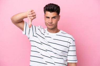 Pembe arka planda izole edilmiş genç beyaz adam olumsuz ifadeyle baş parmağını aşağı gösteriyor.