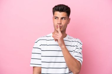 Pembe arka planda izole edilmiş genç beyaz adam sessiz bir jest işareti gösteriyor. Parmağını ağzına sokuyor.