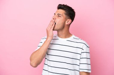 Pembe arka planda izole edilmiş genç beyaz adam esniyor ve ağzını eliyle açık tutuyor.