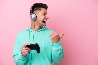 Pembe arka planda izole edilmiş bir bilgisayar oyunu kontrolörüyle oynayan yakışıklı beyaz bir adam bir ürünü sunmak için kenarı işaret ediyor.