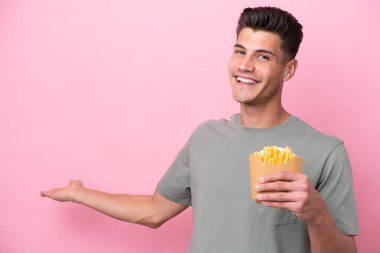 Pembe arka planda izole edilmiş kızarmış cipsleri tutan genç beyaz bir adam davet için ellerini yana uzatıyor.