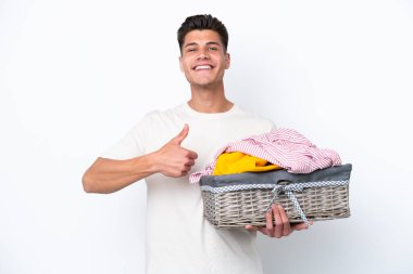 Beyaz arka planda çamaşır sepetini tutan genç beyaz bir adam baş parmak hareketi yapıyor.
