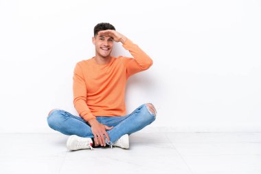 Genç adam beyaz arka planda izole bir şekilde yerde oturuyor. Bir şeye bakmak için uzaklara bakıyor.