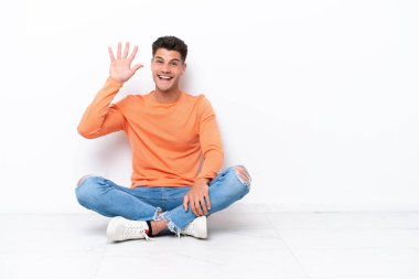 Genç adam beyaz arka planda tek başına oturmuş mutlu bir ifadeyle selamlıyor.