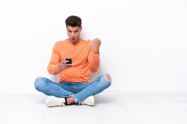 Yerde oturan genç adam beyaz arka planda izole edilmiş, şaşırmış ve bir mesaj gönderiyor.