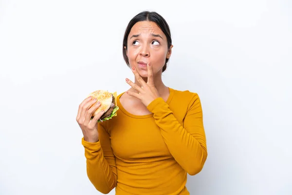 Młoda Latynoska Kobieta Trzyma Hamburgera Białym Tle Mając Wątpliwości Podczas — Zdjęcie stockowe