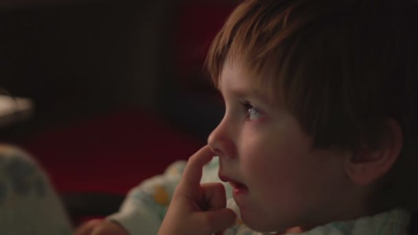 Jonge jongen zijn neus plukken tijdens het kijken naar tekenfilms in een donkere kamer op een computer. — Stockvideo