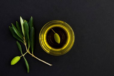Cam bir kasede zeytinyağı ve siyah arka planda yeşil zeytin dalının üst görüntüsü. Boşluğu kopyala.