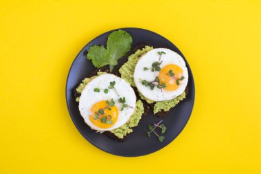 Kahvaltıda çavdar ekmeği, avokado püresi, kızarmış yumurta ve sarı arka planda mikroyeşil ile avokado tostlarının üst görüntüsü.