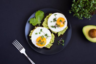 Kahvaltıda çavdar ekmekli avokado tostları, avokado püresi, kızarmış yumurta ve siyah arka planda mikroyeşil. Boşluğu kopyala.