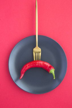 Kırmızı arka plandaki siyah tabakta altın bir çatala saplanmış kırmızı biber. Konum dikey.