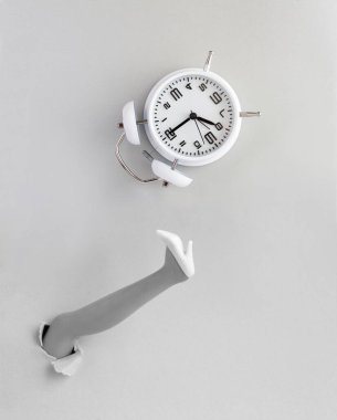 Beyaz ayakkabılı kadın ayağı yırtık bir kağıda sokulmuş ve gri arka planda beyaz alarm saati var. Uzayı kopyala Üst görünüm.