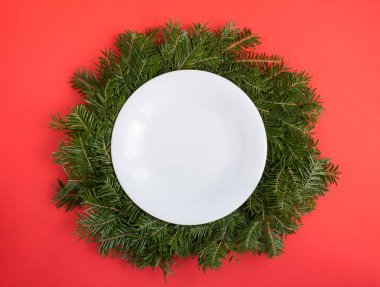 Top view of empty white plate on Christmas ring or wreath on the red background. Copy space.