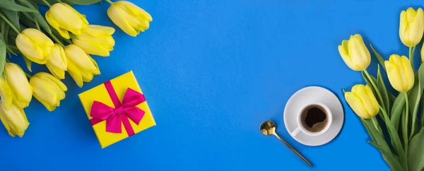 Gift box, yellow tulip and coffee cup on the blue background. Banner. Copy space.