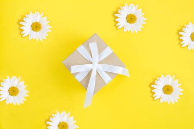 Sarı arka planda bağlı beyaz fiyonklu bir hediye kutusu ve papatya çayı. Üst görünüm.