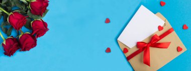 Kırmızı güller, mesaj için boş beyaz kağıt, zarf ve mavi arka planda kırmızı kalp. Uzayı kopyala Tatil afişi.