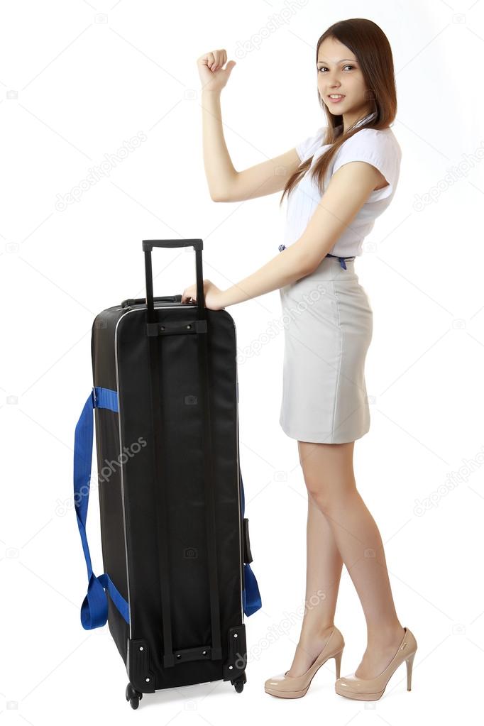 A young, slim girl teenager 16 years old, stands next to a huge, black suitcase on wheels. Girl dressed in a white blouse, high heels and a short skirt. She holds out her hand with a raised thumb.