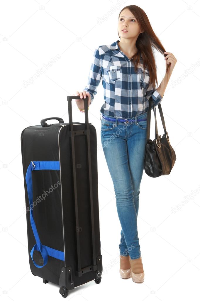 Slim girl in jeans and a plaid shirt, standing next to a big, black travel bag on wheels. The girl pulls hair. One person, teenager, female, vertical image, isolated on white background.