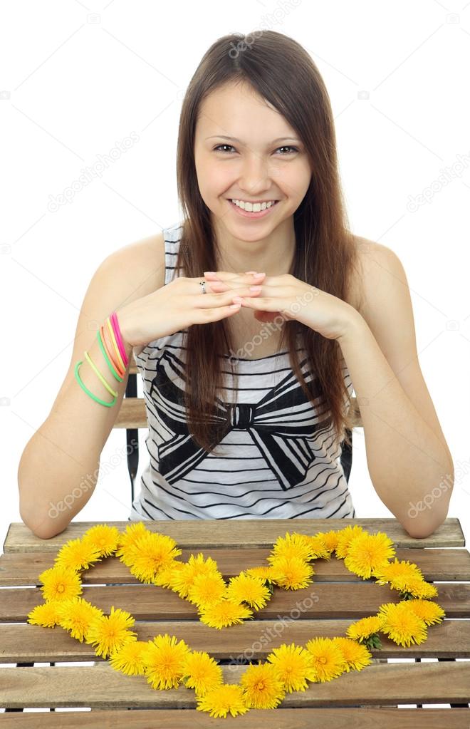 Yellow flowers dandelions in form of Heart. One teenage girl 16 years old, caucasian appearance, laid out on the table the heart of dandelion flowers. One person, female teenager, vertical, isolated.