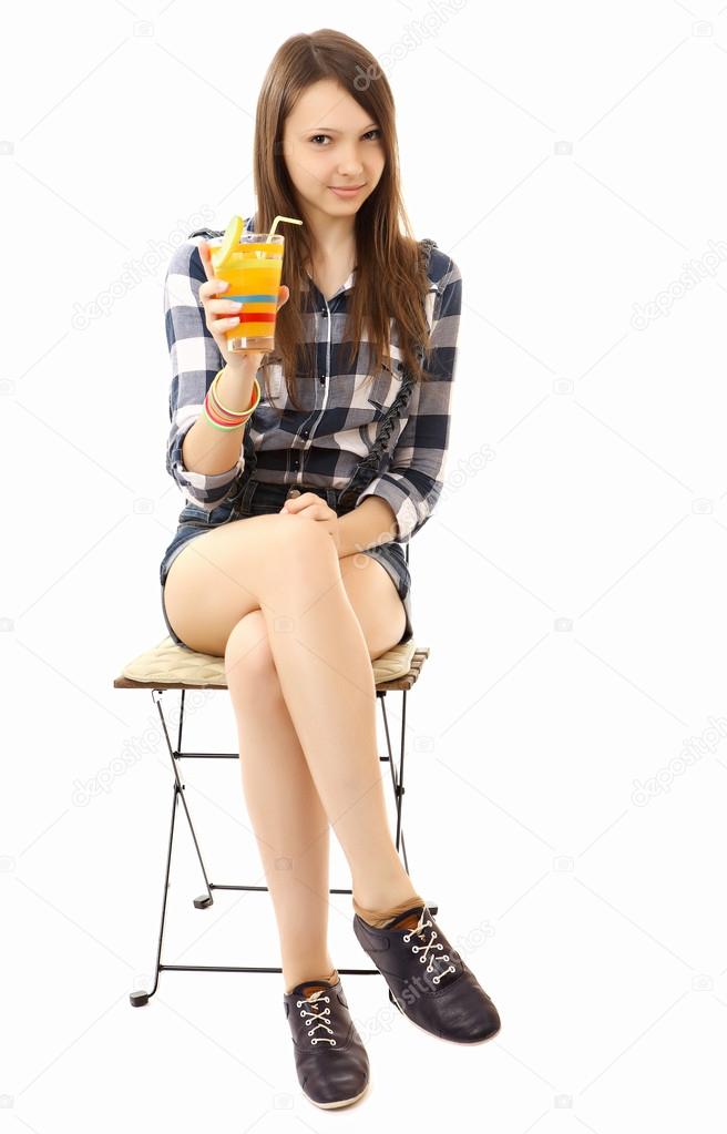 Girl teenager, caucasian appearance, brunette, wearing a plaid shirt and short denim shorts, holding a glass of drink. Teen Girl relaxing sitting on folding chair with a cocktail in hand. One person.