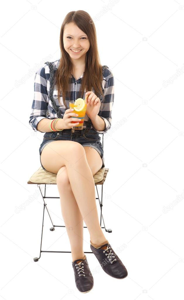Girl teenager, caucasian appearance, brunette, wearing a plaid shirt and short denim shorts, holding a glass of drink. Girl is relaxing, sitting on a folding chair with orange drink in hand, relaxing