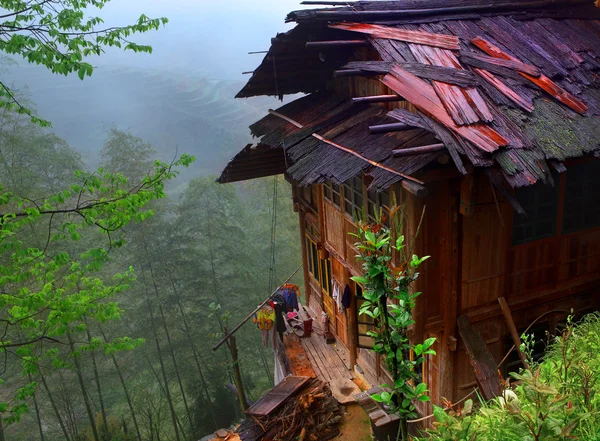 Крестьянский дом с мокрой крышей, стоящий на краю пропасти, с бамбуковыми рощами и рисовыми террасами на заднем плане. Юго-Западный Китай, провинция Гуйчжоу, недалеко от Луншэна. Деревянный дом в горах Китая . — стоковое фото