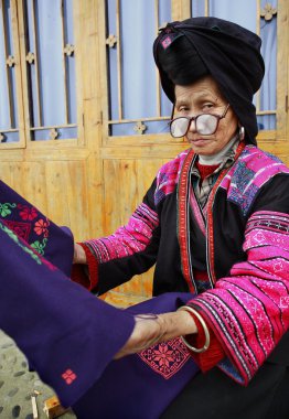 Old woman with big glasses, red Yao nationality, ethnic minorities in China, holding a blue cloth with patterns embroidered by hand, 4 April 2010. Xiaozhai Yao ethnic minority village, near Longsheng clipart