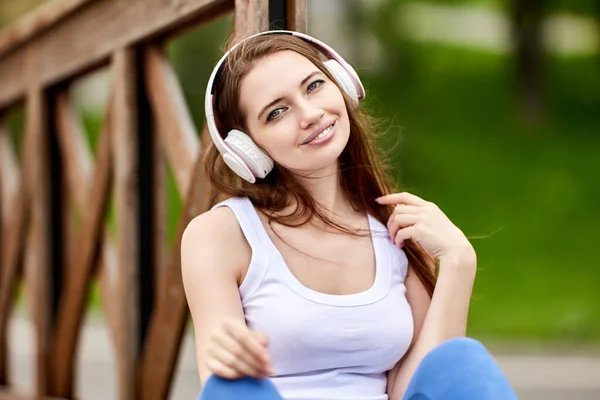 Uśmiechnięta młoda kobieta w słuchawkach bezprzewodowych siedzi na zewnątrz. — Zdjęcie stockowe