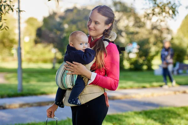 Femme avec bébé en écharpe pour bébé se tient dans le parc public. — Photo