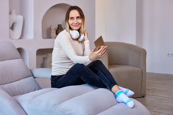 Happy woman in wireless headphones with phone sits on sofa indoors. — Stock Photo, Image