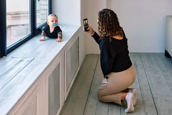 Bébé avec sucette rampe sur le rebord de la fenêtre et la femme prend des photos. — Photo