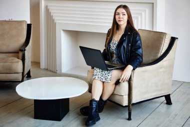 Deri çizmeli ve deri ceketli bir kadın oturma odasında dizüstü bilgisayarla oturuyor..