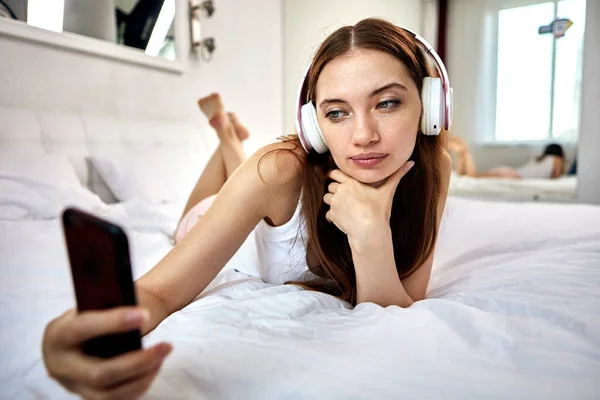 Kobieta w słuchawkach leży w łóżku ze smartfonem. — Zdjęcie stockowe