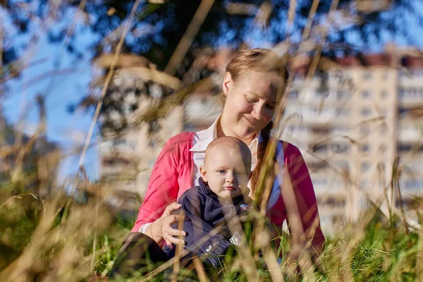 Mother with baby on walk in city park sit in tall grass on summer evening. — Stock Photo, Image